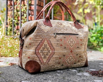 May include: A travel bag featuring a woven fabric body with a tan, brown, and rust-colored geometric pattern. The bag includes brown leather handles, leather corner reinforcements, a top zipper closure, and a horizontal zippered pocket on the front.