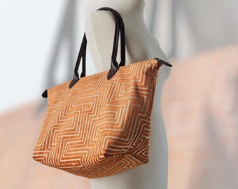 May include: An orange tote bag with a geometric pattern and black leather handles. The bag is displayed on a mannequin. The bag's design features a repeating zig-zag pattern in a lighter shade of orange.