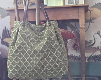May include: A green and white patterned tote bag with brown leather straps. The bag is sitting on a wooden chair in a room with a patterned wallpaper.