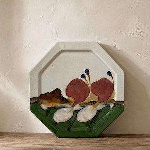 May include: An octagonal ceramic plate with a speckled off-white base. The design features a green band at the bottom, a brown mountain-like shape, and two large red floral-like forms with thin stems and small blue circular accents at the top.