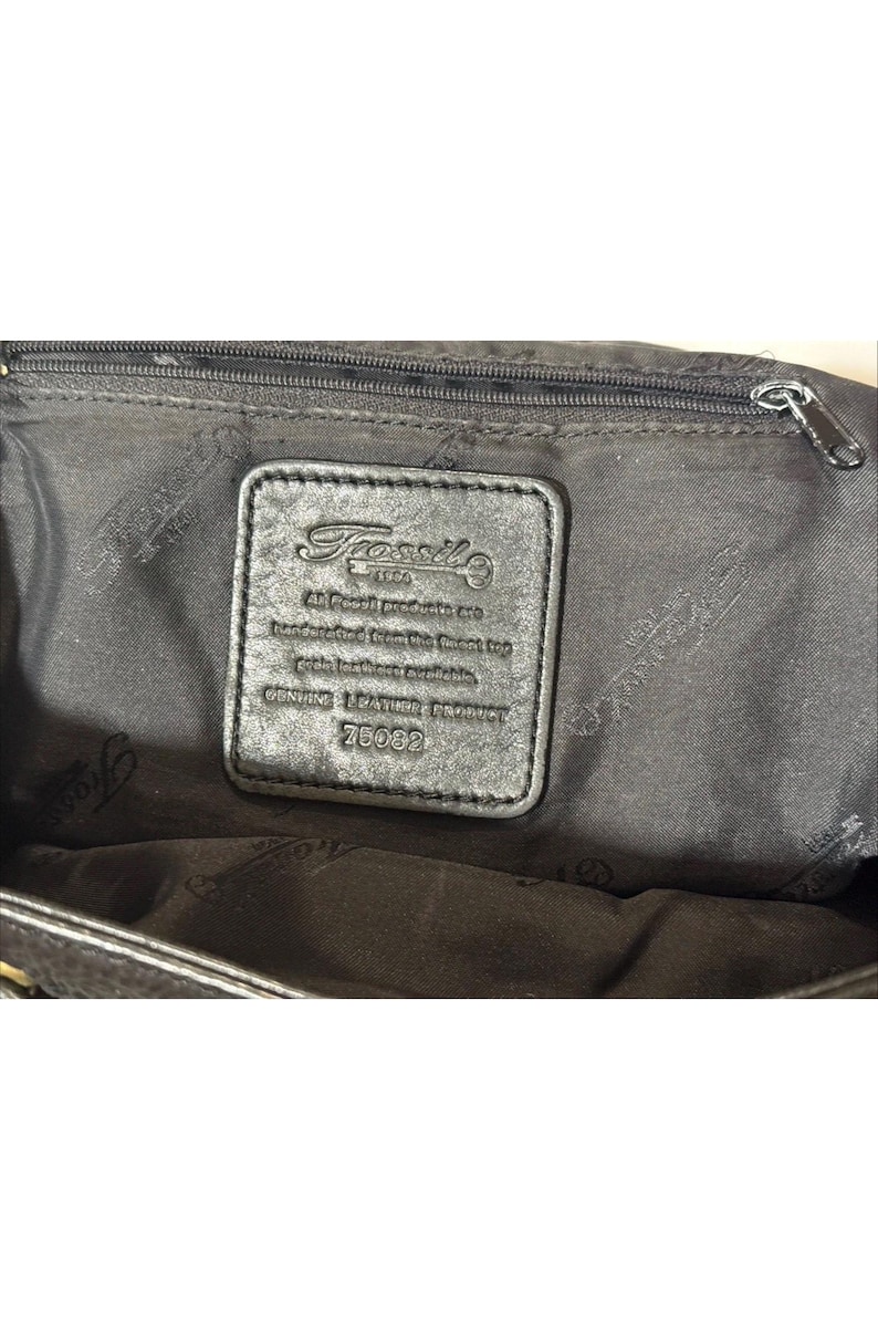May include: Black leather handbag interior with a zipper and a rectangular leather tag. The tag features the Fossil logo and text: "All Fossil products are handcrafted from the finest top grain leathers available. Genuine leather product 76082."