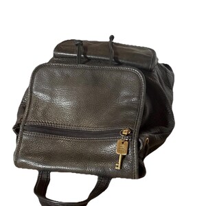 May include: A black leather backpack with a top flap and a front zippered pocket. The backpack has a small handle and adjustable straps. A gold-colored key hangs from the zipper. The leather has a textured appearance.