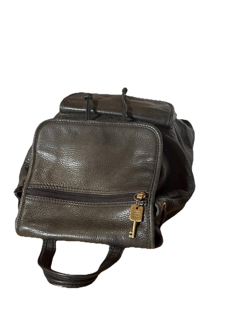 May include: A black leather backpack with a top flap and a front zippered pocket. The backpack has a small handle and adjustable straps. A gold-colored key hangs from the zipper. The leather has a textured appearance.