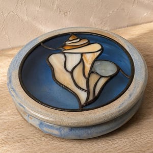 May include: A round ceramic lidded box featuring a stained glass inlay on the top. The inlay depicts a cream and tan seashell against a deep blue background, framed by dark metal lines. The box has a speckled, light blue and beige glazed finish.