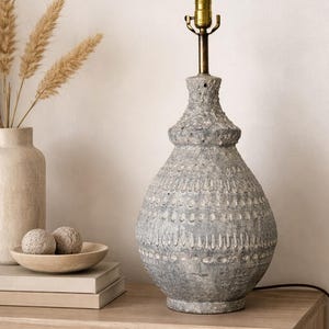 May include: A gray ceramic table lamp with a textured surface and brass hardware. The lamp has a bulbous base and a decorative pattern. It sits on a wooden surface, next to a vase and decorative spheres.