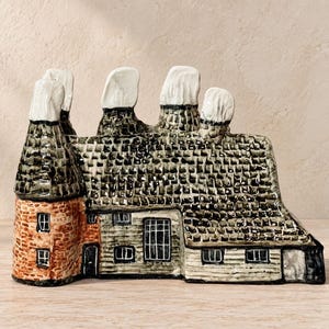 May include: A ceramic house figurine featuring a round brick-patterned tower, a dark shingled roof, and four white chimney stacks. The structure includes small rectangular windows and a dark door, with a light-colored base and walls.