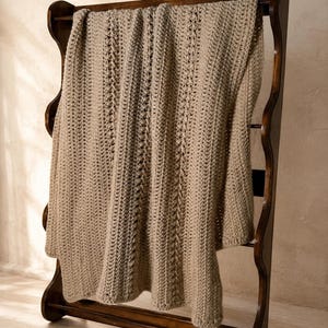 May include: A beige crochet blanket with a textured vertical pattern featuring alternating rows of solid stitches and raised cable-like details. The blanket is draped over a dark brown wooden rack with curved side supports and horizontal bars.