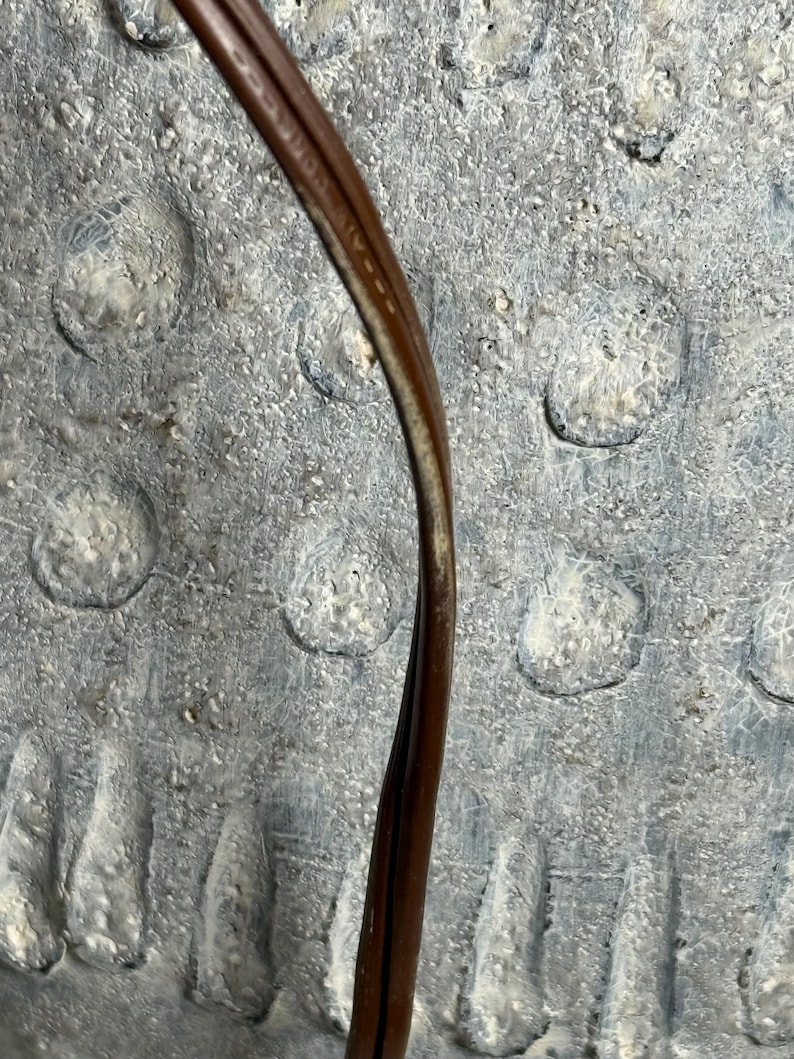 May include: A close-up of a brown electrical cord against a textured, gray background. The cord is curved and appears to be made of a flexible material. The background has a mottled appearance with circular indentations.