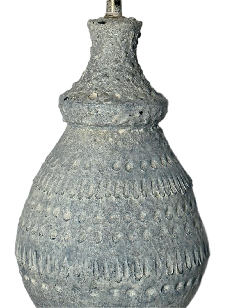 May include: A textured, gray ceramic lamp base with a bulbous shape and a detailed, layered design. The lamp base has a rough, pitted surface and a silver-colored metal fitting at the top. The overall aesthetic is rustic and artisanal.