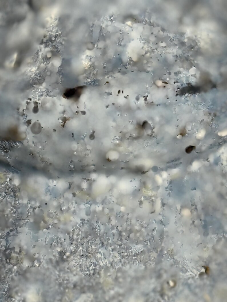 May include: Close-up of a textured, abstract surface with a mix of light blue, white, and gray tones. The surface has a rough, uneven appearance with small, dark spots and a bubbly texture, creating a unique visual effect.