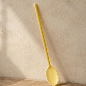 May include: A pale yellow spoon with a long handle, leaning against a textured wall. The spoon has an oval-shaped bowl with the word "Splendor" embossed on it. The spoon is made of a solid, smooth material.