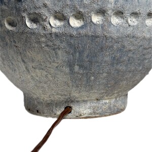 May include: Close-up of a textured, blue-gray ceramic lamp base. The lamp base has a rounded shape with a series of circular indentations and vertical grooves. A brown electrical cord extends from the base.