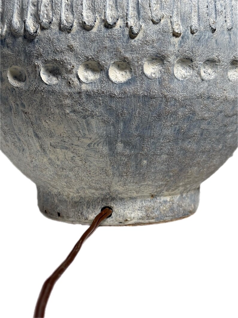 May include: Close-up of a textured, blue-gray ceramic lamp base. The lamp base has a rounded shape with a series of circular indentations and vertical grooves. A brown electrical cord extends from the base.