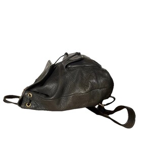 May include: Black leather drawstring backpack with a textured surface. The bag features gold-colored eyelets and adjustable shoulder straps. The bag is designed with a top flap and a drawstring closure.