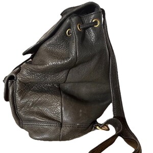 May include: Black leather backpack with a drawstring closure and flap. The bag features a textured surface, gold-toned hardware, and adjustable shoulder straps. A small pocket is visible on the front.