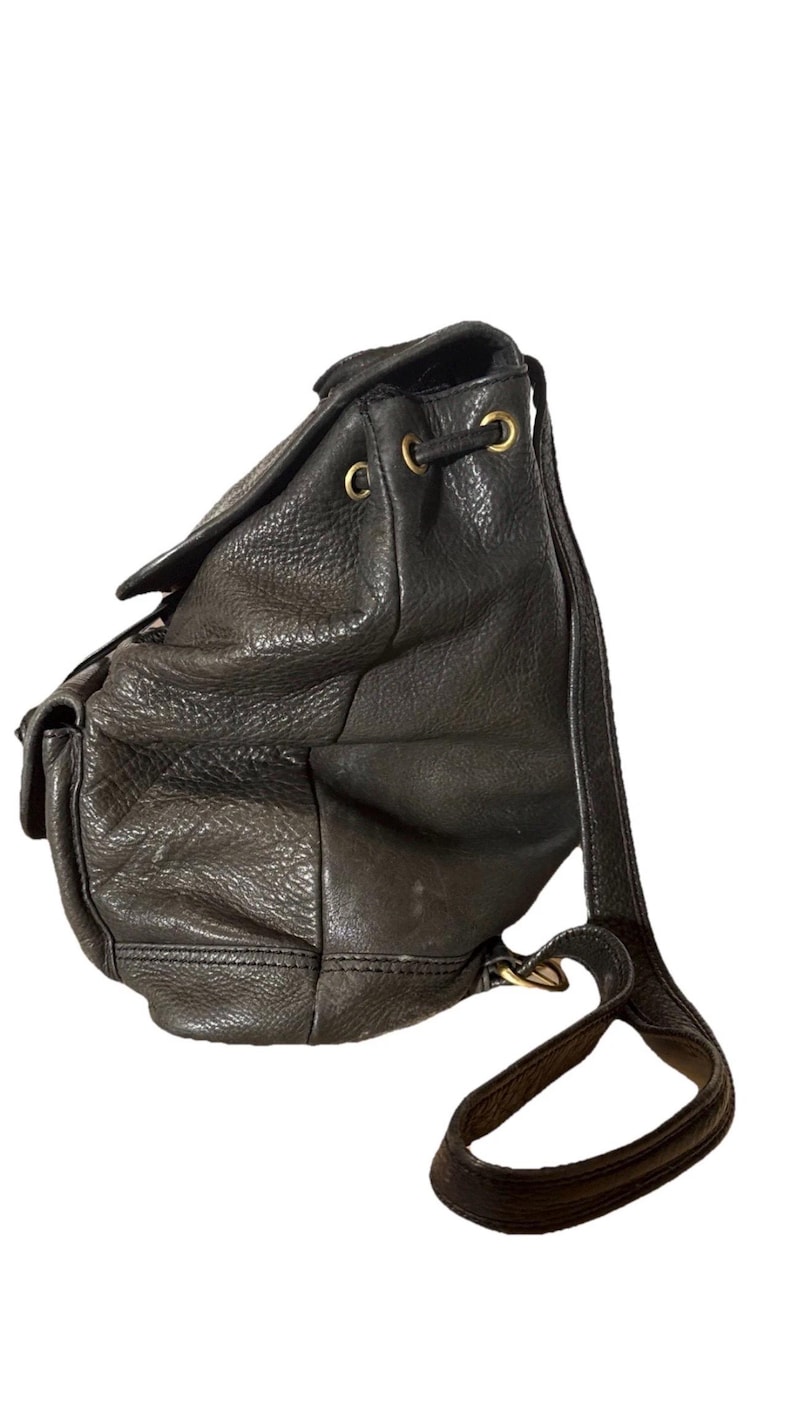 May include: Black leather backpack with a drawstring closure and flap. The bag features a textured surface, gold-toned hardware, and adjustable shoulder straps. A small pocket is visible on the front.