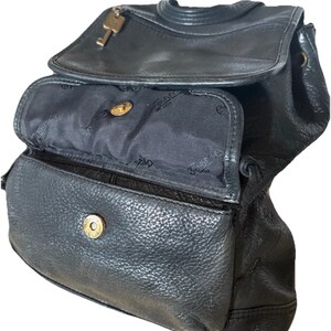 May include: Black leather backpack with a flap closure and a key charm. The bag has multiple compartments, including a front pocket and a main compartment with a snap closure. The interior lining is black.
