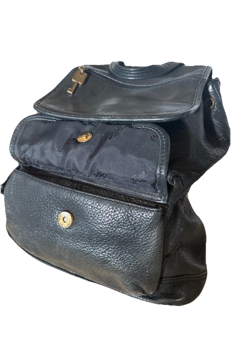 May include: Black leather backpack with a flap closure and a key charm. The bag has multiple compartments, including a front pocket and a main compartment with a snap closure. The interior lining is black.