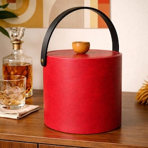 May include: A red ice bucket with a black handle and a wooden lid. The cylindrical bucket has a black handle and a small wooden knob on top. The bucket is sitting on a wooden surface, with a decanter and glass in the background.