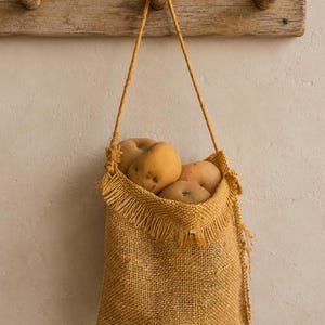 May include: A rustic, mustard-colored burlap bag filled with several light brown potatoes, hanging from a wooden rack. The bag has a fringed top and a woven texture, suggesting a natural, handmade aesthetic.