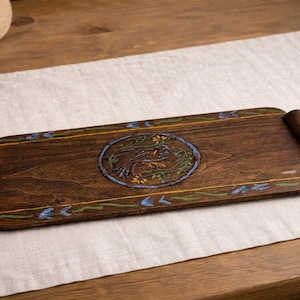 May include: A rectangular wooden serving tray with dark brown stain and rolled handles. The tray features a central oval design with a bird and floral motif in blue, yellow, and green. The edges are decorated with painted floral accents.