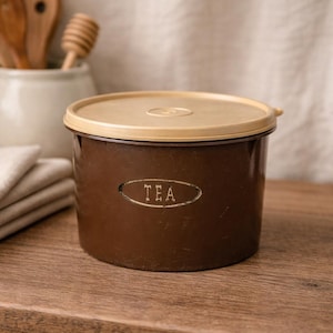 May include: A brown tea canister with a tan lid. The word "TEA" is printed on the front in a white, oval-shaped frame. The canister is sitting on a wooden surface.