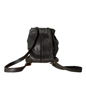 May include: Black leather backpack with adjustable straps and a zippered main compartment. The backpack has a textured finish and brass-colored hardware. The straps are attached with brass buckles. The bag is designed to be worn on the back.