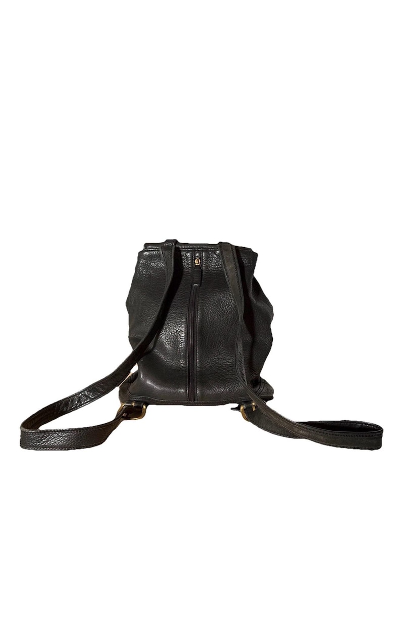 May include: Black leather backpack with adjustable straps and a zippered main compartment. The backpack has a textured finish and brass-colored hardware. The straps are attached with brass buckles. The bag is designed to be worn on the back.