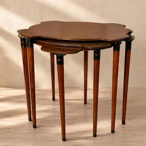 May include: A set of three nested tables with dark brown wooden tops featuring scalloped edges. Each table has four slender, tapered legs in a matching brown finish, accented with black and gold horizontal bands near the top of each leg.