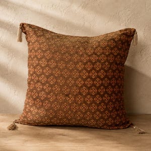 May include: A decorative square pillow with a repeating floral pattern in shades of brown and orange. The pillow has tassels at each corner and is placed on a wooden surface.