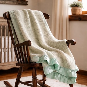 May include: A wooden rocking chair with a light green knitted blanket draped over it. The blanket has a ruffled, light blue trim. The chair is on a white rug, and the background includes a crib and a window.