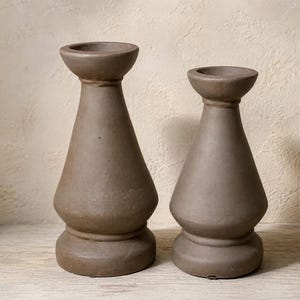 May include: Two brown candle holders with a matte finish, featuring a wide circular base, a tapered body, and a flared top rim. The candle holders are constructed with a smooth, solid surface and stand upright, displaying a consistent earthy tone.