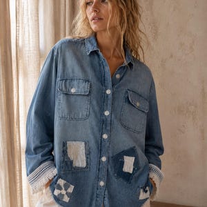 May include: A light blue denim shirt with long sleeves, two chest pockets, and white buttons. The shirt features decorative patches and lace trim on the cuffs. The model is wearing white pants.
