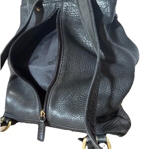 May include: Black leather backpack with a textured finish. The bag features a zippered front pocket and adjustable straps. Gold-colored hardware accents the design, including the zippers and strap attachments. The interior lining is visible through the open zipper.