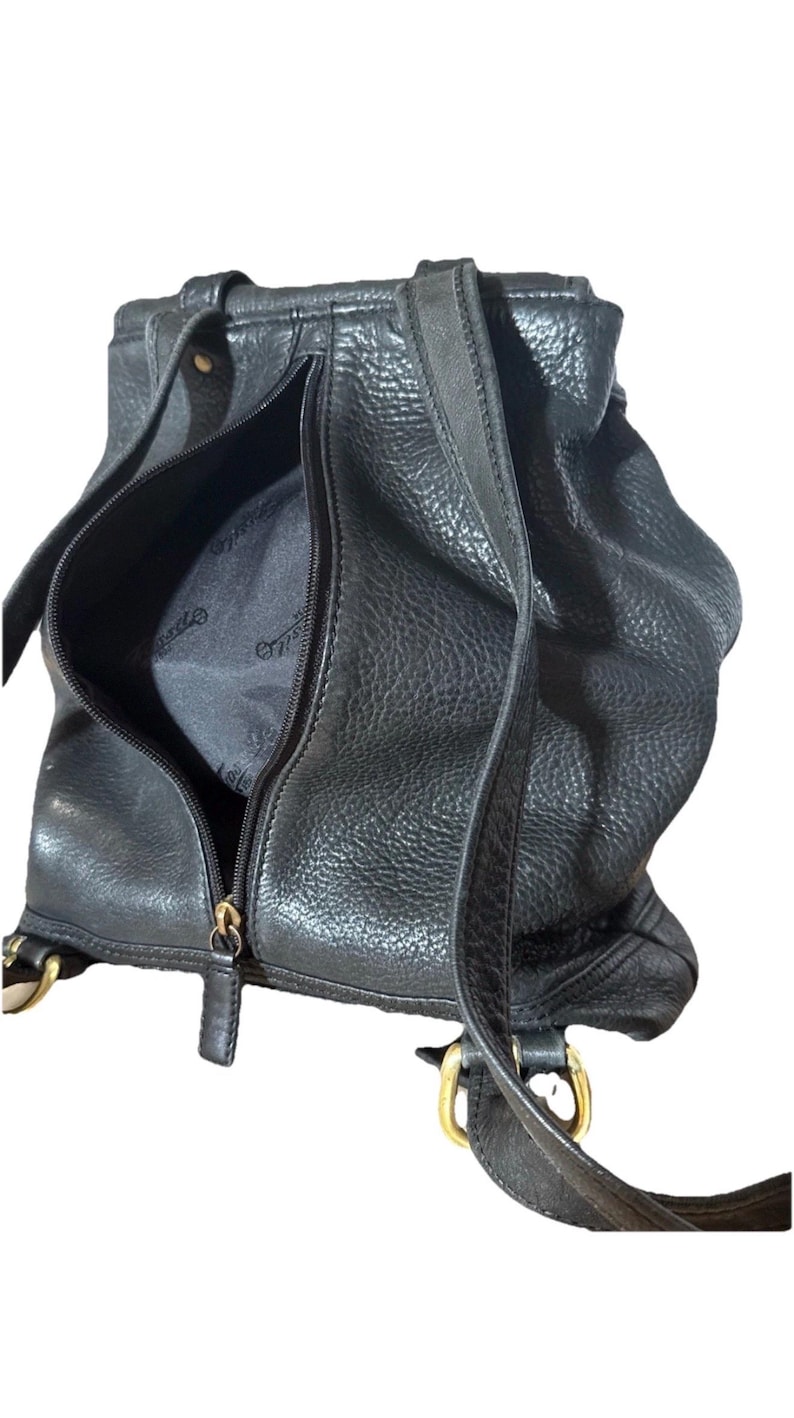 May include: Black leather backpack with a textured finish. The bag features a zippered front pocket and adjustable straps. Gold-colored hardware accents the design, including the zippers and strap attachments. The interior lining is visible through the open zipper.
