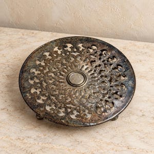 May include: A round, silver-toned metal trivet featuring an intricate, openwork filigree pattern of swirling leaves and vines. The surface displays a darkened, aged patina with a smooth, circular center medallion and small, sturdy feet.