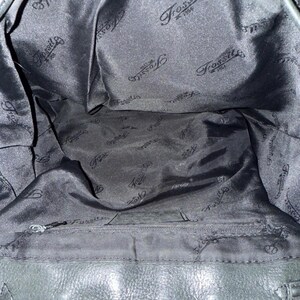 May include: Interior view of a black Fossil handbag. The lining is black satin with the Fossil logo printed repeatedly. A zippered pocket is visible at the bottom. The bag's leather exterior is also black.