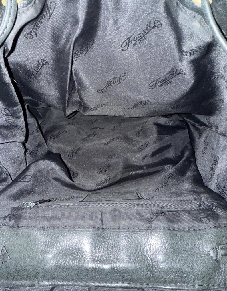 May include: Interior view of a black Fossil handbag. The lining is black satin with the Fossil logo printed repeatedly. A zippered pocket is visible at the bottom. The bag's leather exterior is also black.