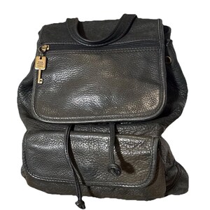 May include: Black leather backpack with a flap closure and a drawstring. The backpack has a top handle, a zippered pocket, and a key-shaped charm. The bag has a textured surface and a classic design.
