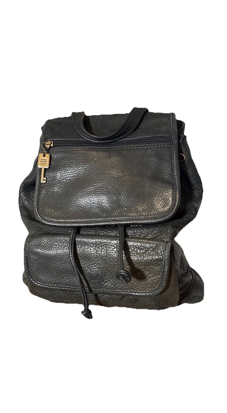 May include: Black leather backpack with a flap closure and a drawstring. The backpack has a top handle, a zippered pocket, and a key-shaped charm. The bag has a textured surface and a classic design.