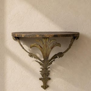 May include: A wall-mounted console shelf featuring a semi-circular top with a dark, mottled brown and gold finish. The shelf is supported by a decorative metal bracket shaped like stylized acanthus leaves in a muted, weathered olive green tone.
