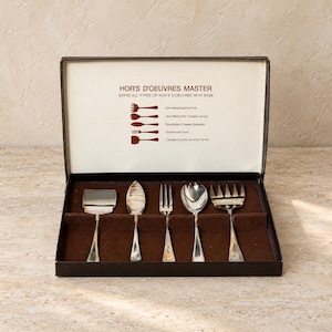 May include: A silver-toned hors d'oeuvres serving set in a black box. The lid interior features the text HOR'S D'OEUVRES MASTER and diagrams of five utensils: a sardine fork, cheese scoop, spreader, pickle fork, and quiche turner.
