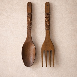 May include: A wooden spoon and fork set with carved handles. The spoon has a rounded bowl, and the fork has three tines. Both utensils are a warm brown color and are hanging on a light beige wall.