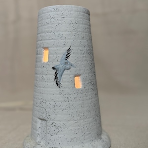May include: A ceramic lighthouse-shaped candle holder. The gray and white speckled holder features a flying seagull design and rectangular cutouts that emit a warm glow. The base is round and the top is open.