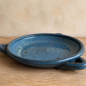 May include: A round, blue ceramic dish with a glossy, mottled glaze finish. The dish features two integrated handles on opposite sides and a series of small, evenly spaced drainage holes visible across the flat interior base of the vessel.