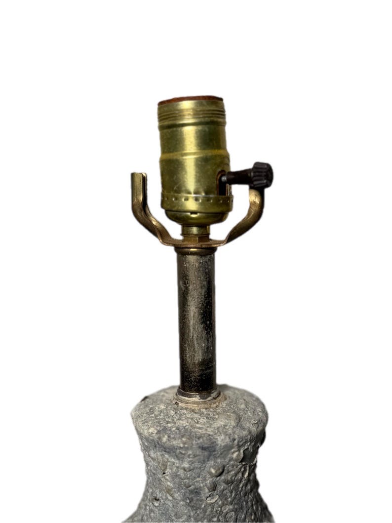 May include: A vintage lamp base with a textured, gray stone-like appearance. The lamp features a brass-colored socket and a black switch. The lamp is designed to hold a light bulb and provide illumination.