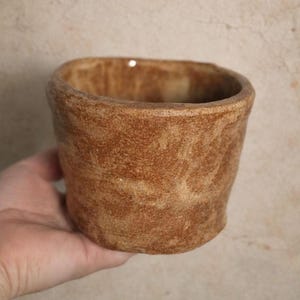 May include: A handmade, brown ceramic cup with a textured surface. The cup has a rounded rectangular shape and a slightly flared rim. The exterior features variations in color, giving it a rustic appearance. The cup is held in a hand.