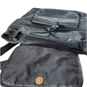 May include: Black leather backpack with a drawstring closure and a front pocket. The interior lining features a repeating "Fossil" logo pattern. A gold-colored button secures the flap.