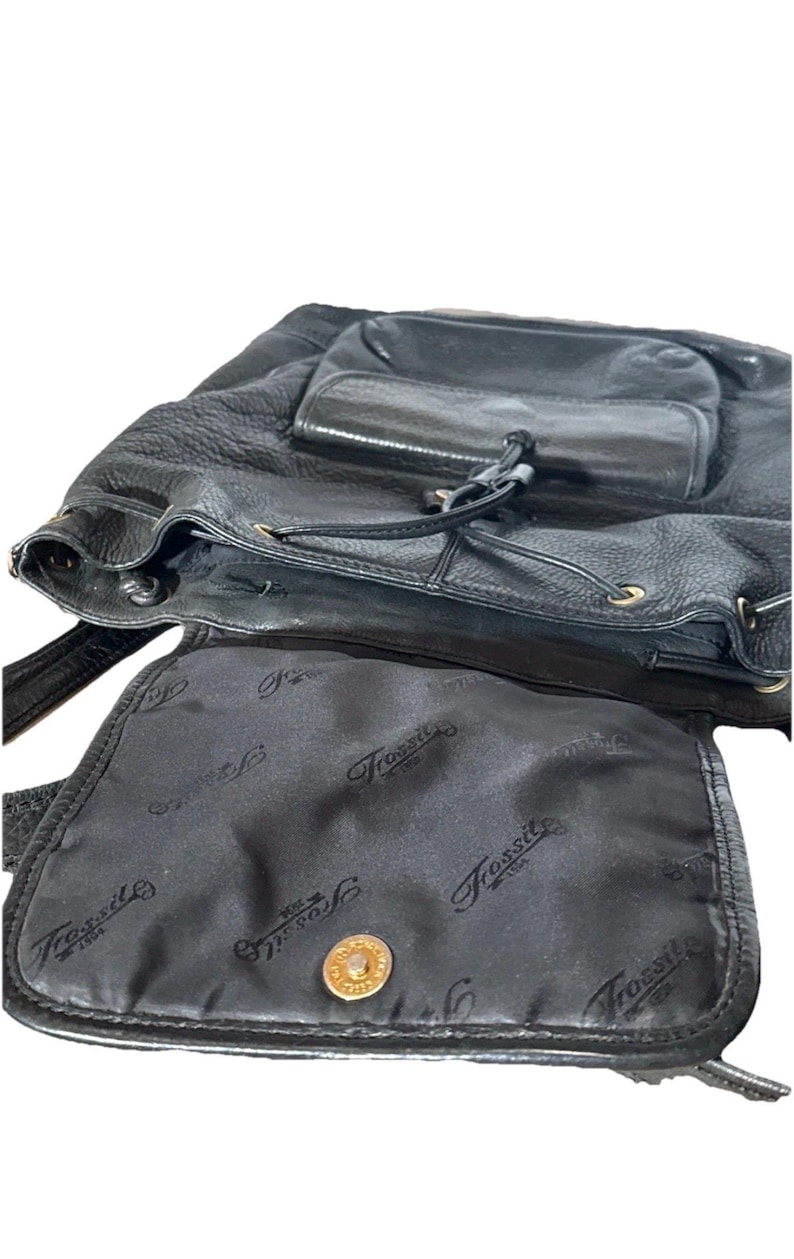 May include: Black leather backpack with a drawstring closure and a front pocket. The interior lining features a repeating "Fossil" logo pattern. A gold-colored button secures the flap.