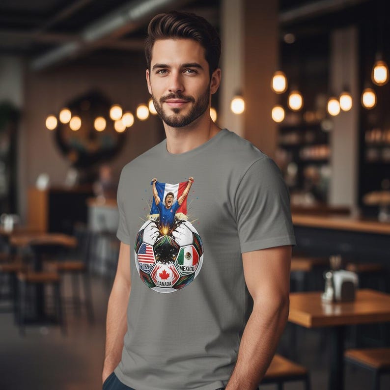 Op de afbeelding: Een grijs t-shirt met een voetbal met de vlaggen van de VS, Canada en Mexico. Een persoon met een Franse vlag staat bovenop de bal. Het t-shirt wordt door een man gedragen.